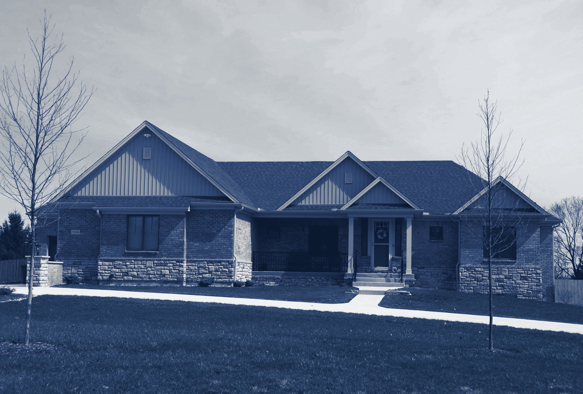 Finished single-story brick-and-stone family home with front porch, gables, young trees and a green lawn, tinted navy