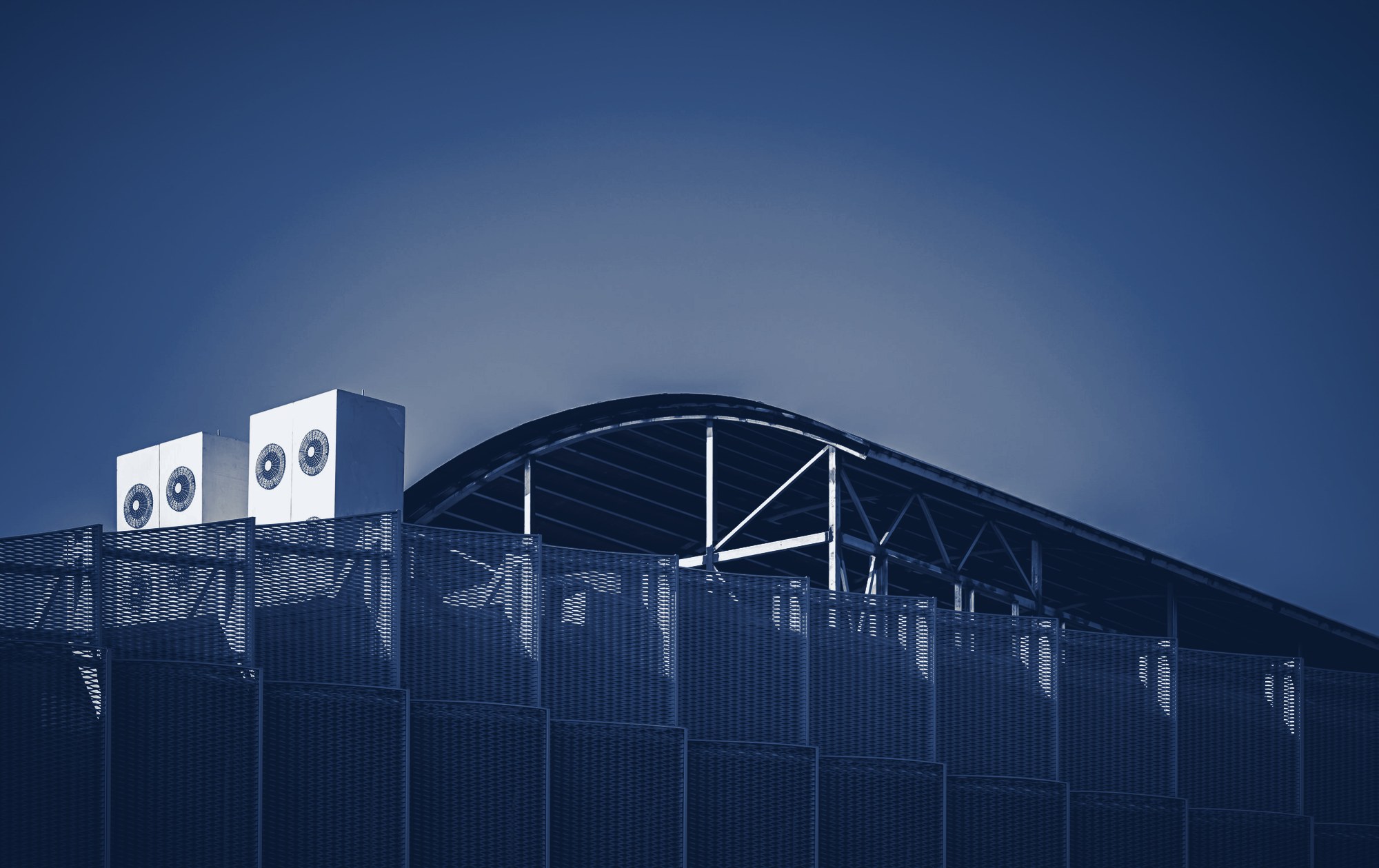 Rooftop HVAC/condenser units behind perforated metal screening on a building with an arched roof, clear blue sky, tinted navy