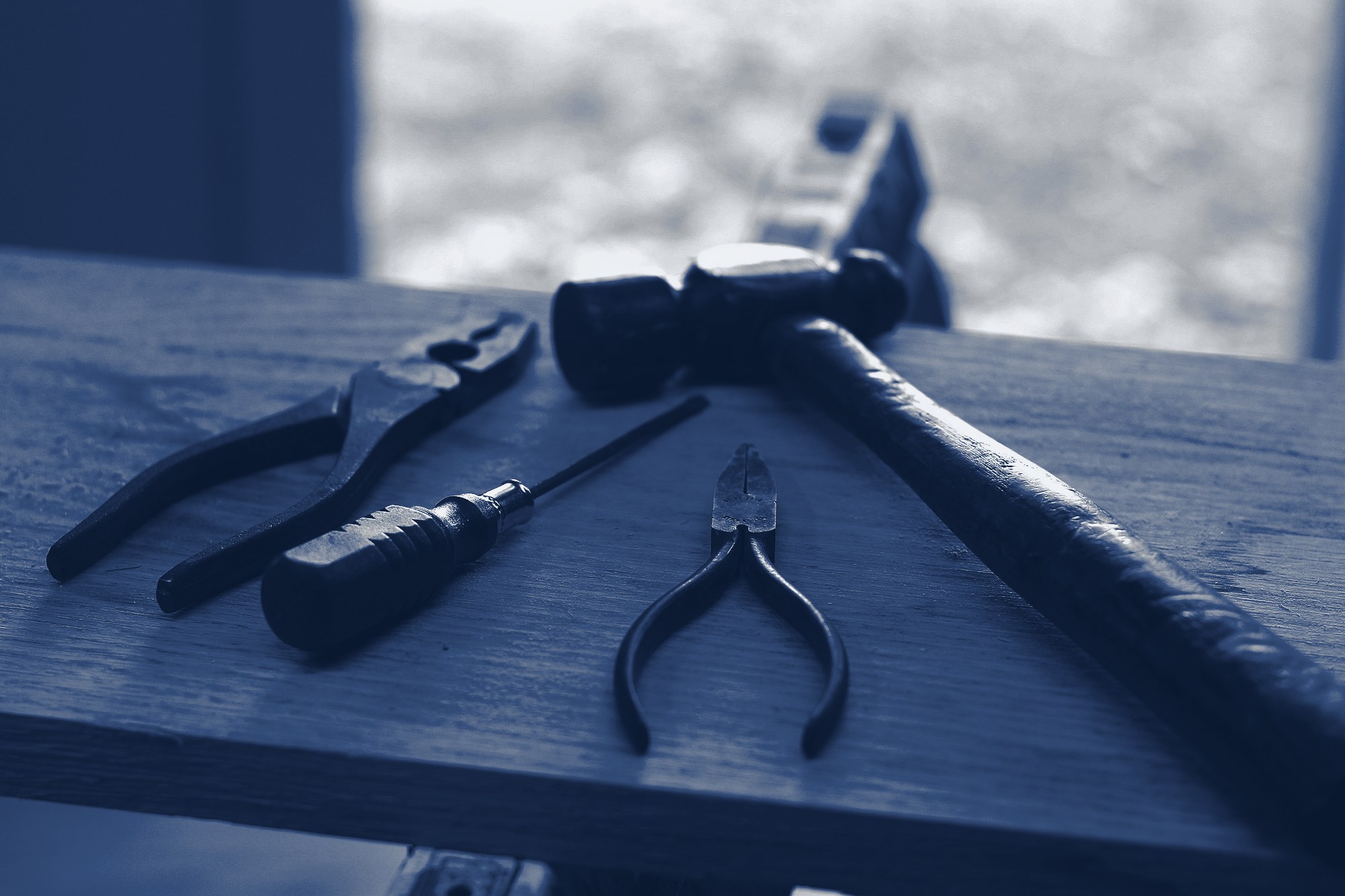Close-up of maintenance hand tools (hammer, pliers, screwdriver) on a wooden board, tinted navy